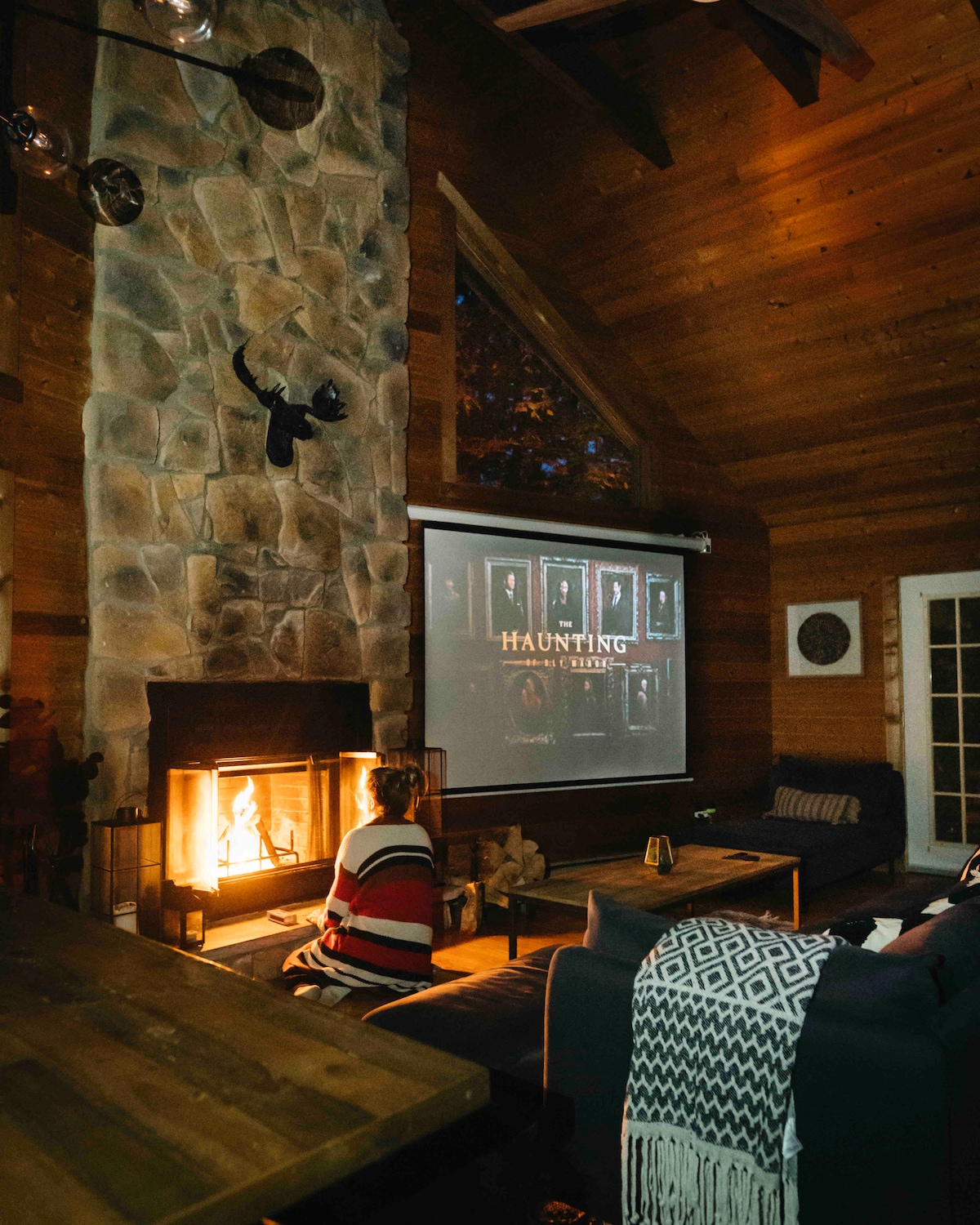 A spacious living room features a stone fireplace, providing warmth and ambiance. A large projector screen displays a movie against the wooden walls. A seating area is arranged with comfortable couches and a knitted throw, inviting relaxation in this cozy setting.