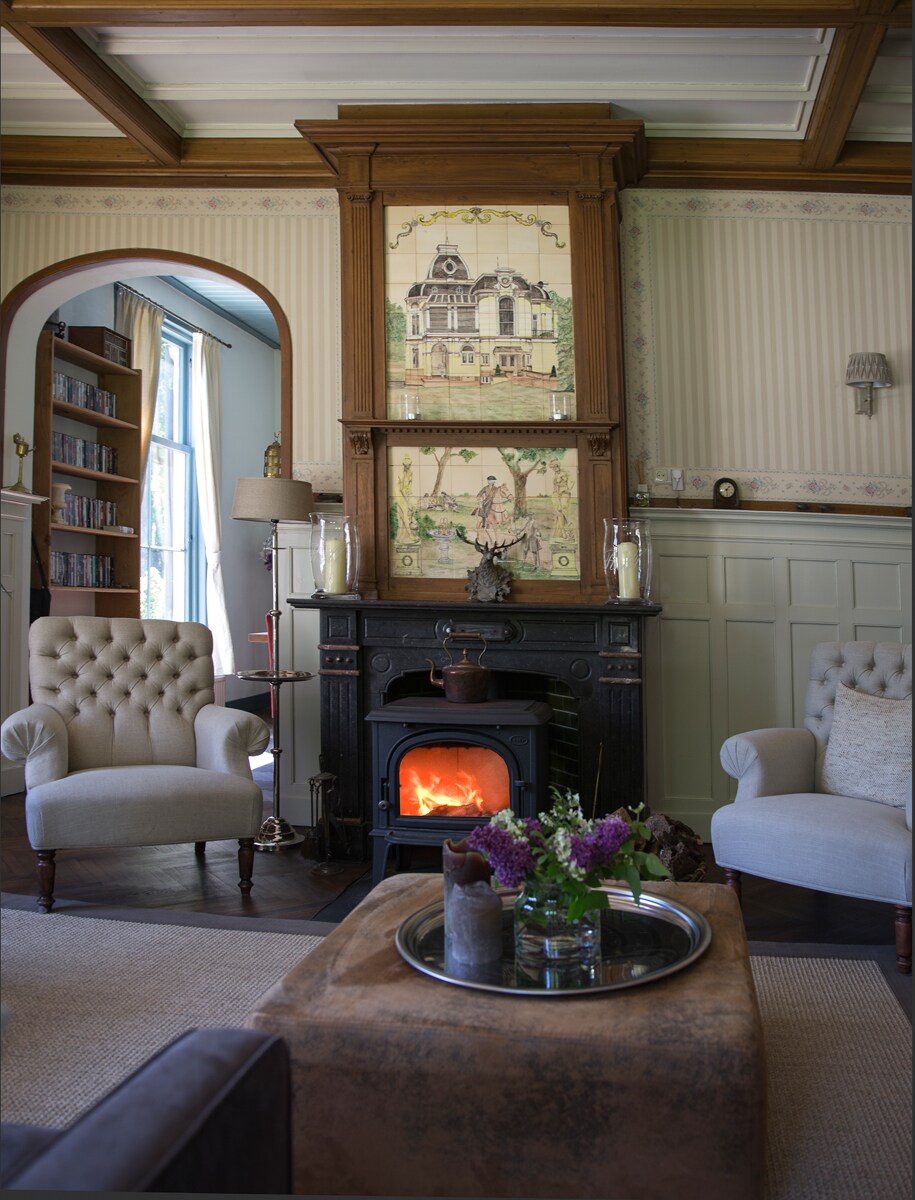 The cozy lounge features two upholstered armchairs positioned beside a traditional wood stove, which emits a soft glow. A decorative fireplace mantel showcases intricate artwork, while a coffee table with a silver tray adds a stylish touch. Natural light streams in through a nearby window.