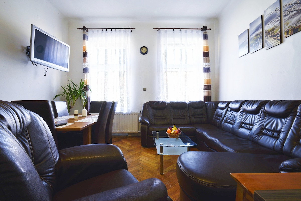 A spacious living room showcases a large, dark leather sectional sofa arranged around a glass coffee table. Natural light filters through two tall windows, accentuated by light curtains. A flat-screen TV is mounted on the wall, while a dining table for four is positioned nearby.