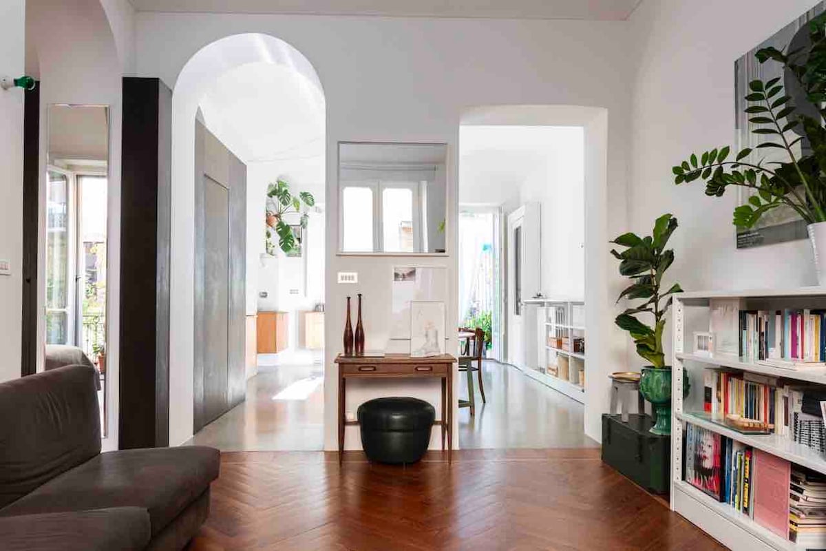 A spacious living area features a comfortable sofa and a small table, with light wood flooring enhancing the room's openness. Plants are visible through an archway, leading toward a bright kitchen area. A bookshelf displays neatly arranged books, contributing to a contemporary aesthetic.