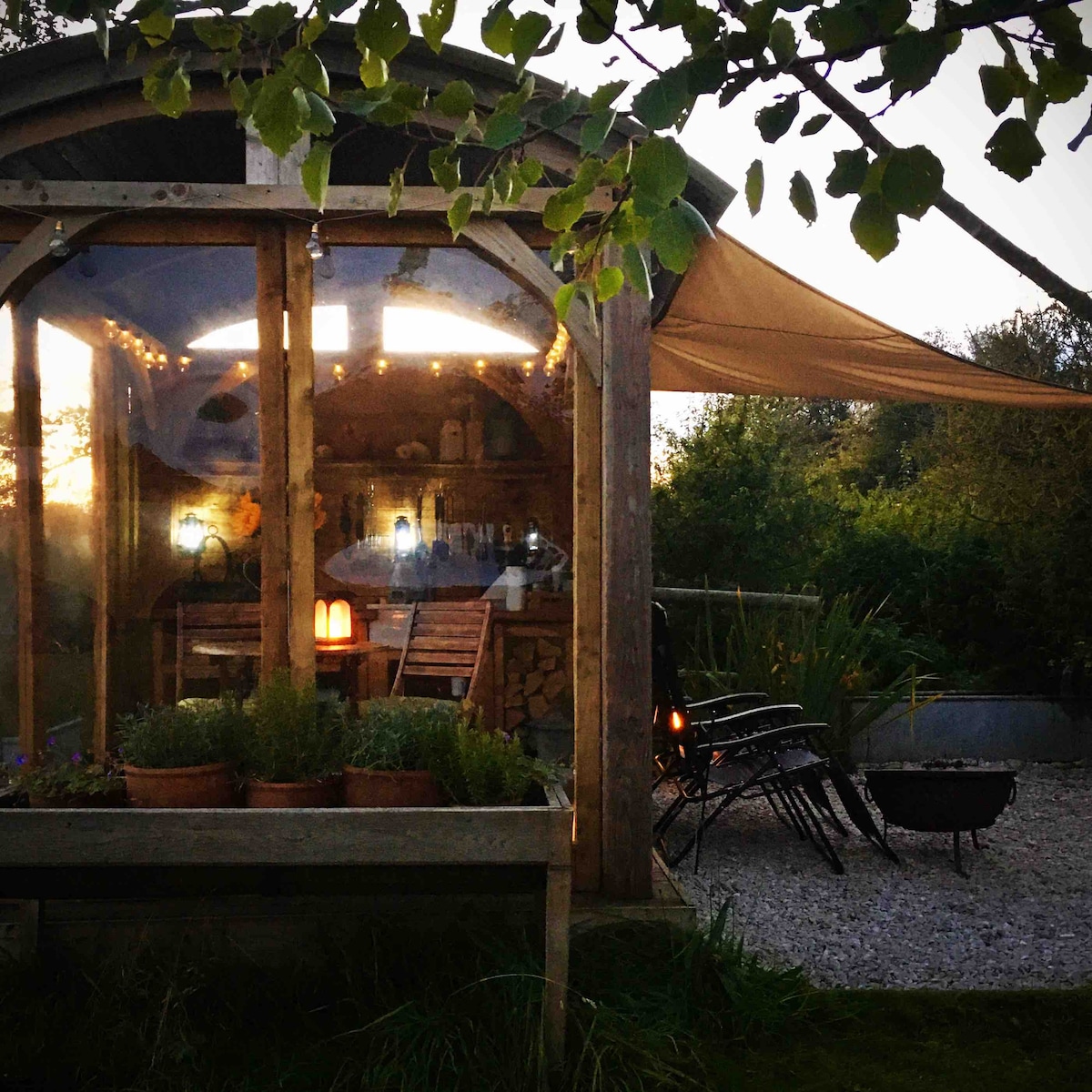 The shepherd's hut is illuminated by soft festoon lights at dusk, featuring large glass windows that offer glimpses of the cozy interior. Outdoor seating surrounds a fire pit, with planters filled with lavender enhancing the tranquil setting. A shaded area provides a welcoming outdoor space.
