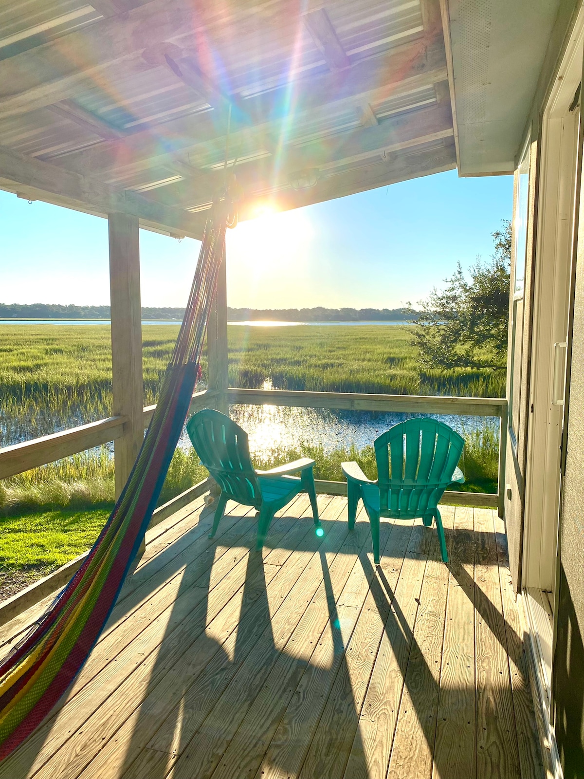 A serene outdoor space features two green adirondack chairs alongside a colorful hammock draped between the posts. Gentle sunlight filters through the scene, illuminating the wooden deck and casting soft shadows on the flooring. The marshland and water views are visible in the distance.