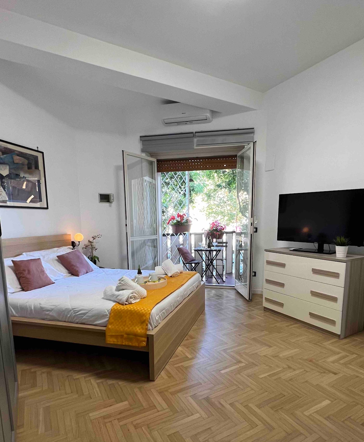 A bright bedroom features a spacious bed with crisp white linens and soft throw pillows. Balcony doors open to a greenery view, and a flat-screen TV is mounted beside a light-colored dresser. Natural light enhances the warm wooden flooring.