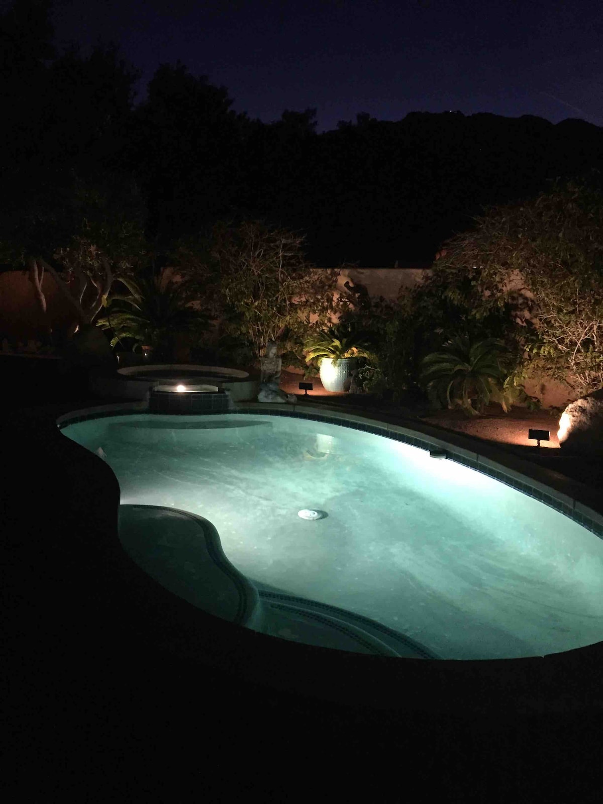 A private swimming pool is illuminated softly at night, surrounded by lush greenery. The serene water reflects gentle lighting from fixtures above, creating a peaceful atmosphere. A hot tub is positioned nearby, contributing to the tranquil setting.