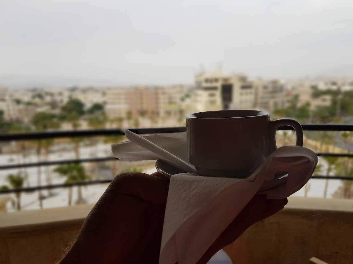 A cup of coffee is held on a saucer, accompanied by a napkin. The background reveals a cityscape with a mix of buildings and greenery, under a soft gray sky.