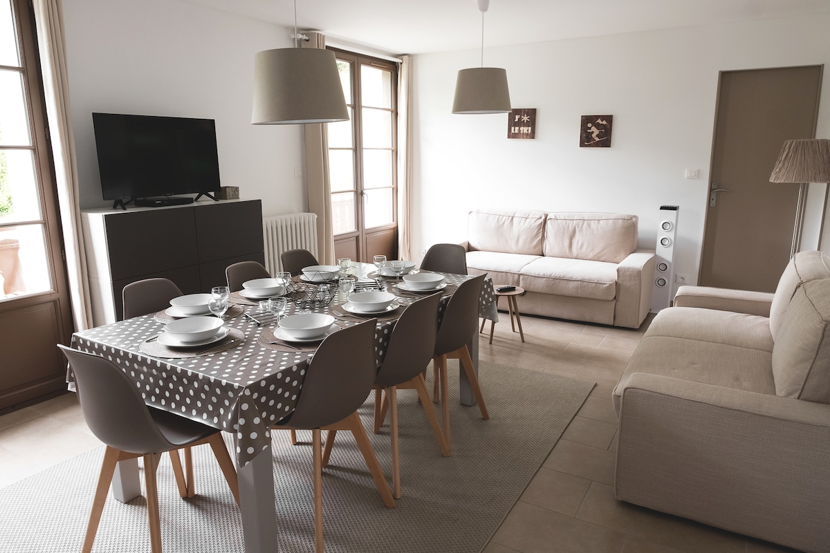 A modern dining area features a large table set for eight with a polka-dot tablecloth. A light-filled space is complemented by a comfortable sofa and a television. Large windows allow for ample natural light, creating a spacious environment.