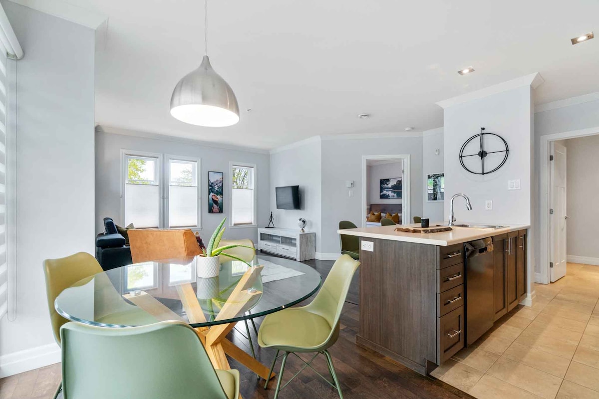 A modern open-concept living space is depicted, featuring a glass dining table surrounded by green chairs. An island with cabinets and a stovetop is visible in the kitchen area. Comfortable seating and a television are present in the background, with natural light illuminating the room.