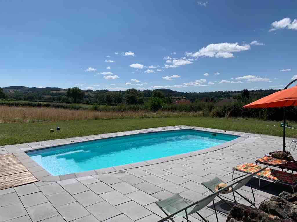 A clear swimming pool is surrounded by a tiled patio, offering sun loungers and an umbrella for shade. The expansive green landscape extends in the background, with rolling hills and a bright blue sky dotted with fluffy clouds.