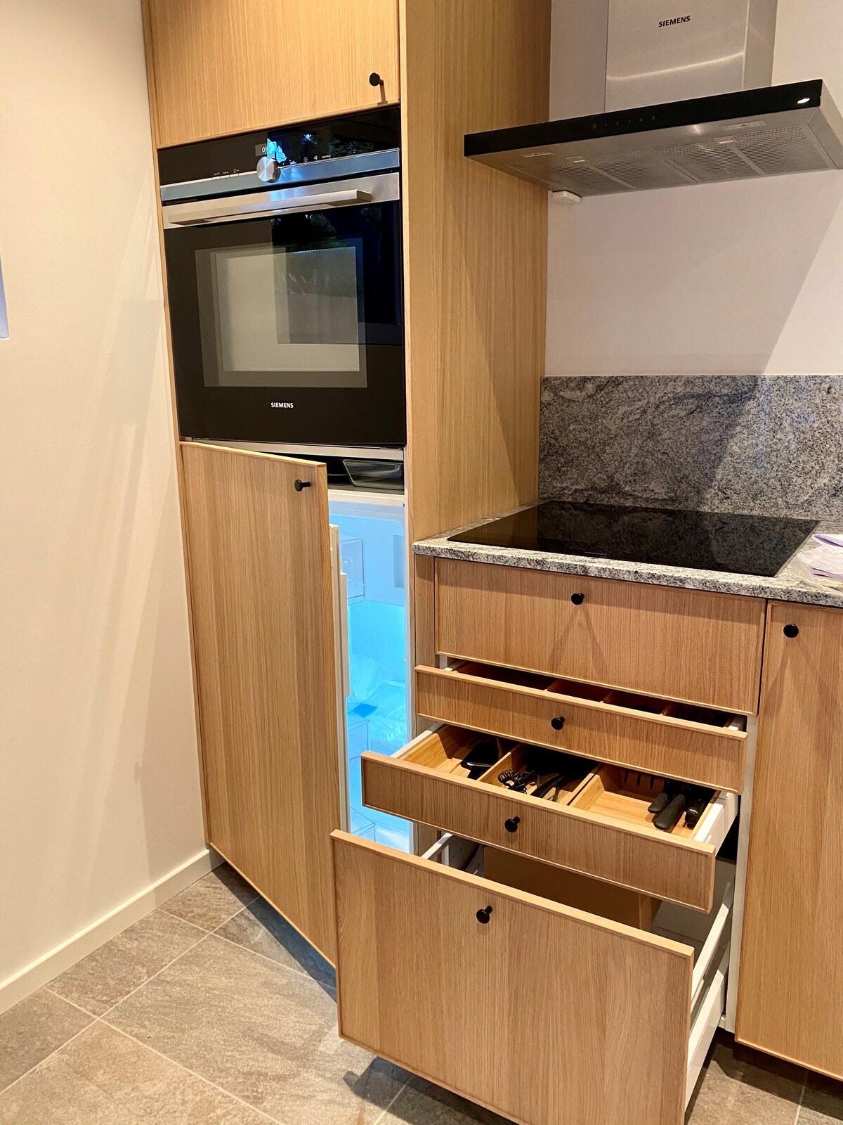 The kitchen features modern wooden cabinetry with a black oven and a granite countertop. A well-organized design is indicated by open drawers displaying utensils. An illuminated refrigerator is integrated into the cabinetry, providing easy access to food storage.
