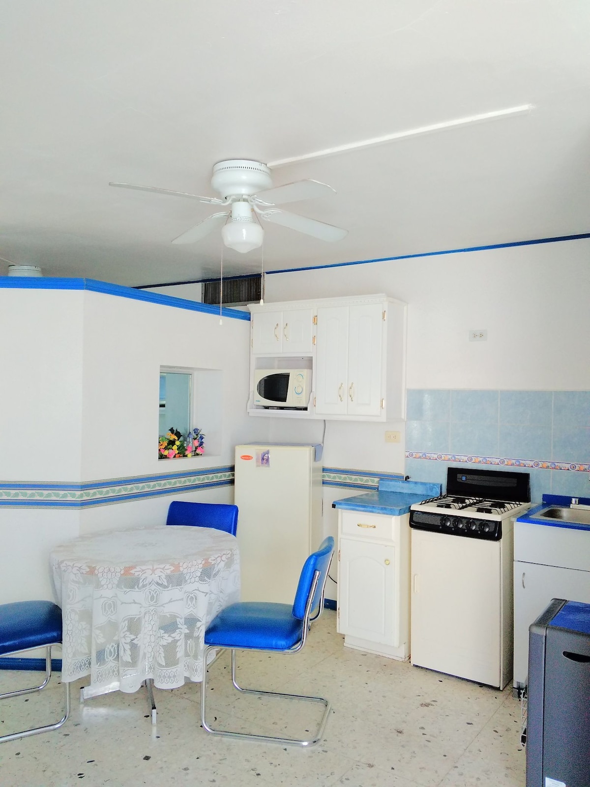The kitchen area features a round dining table with four blue chairs. A ceiling fan provides ventilation, while essential appliances, including a refrigerator, stove, and microwave, are neatly arranged against the walls. Light-colored cabinets offer ample storage space.