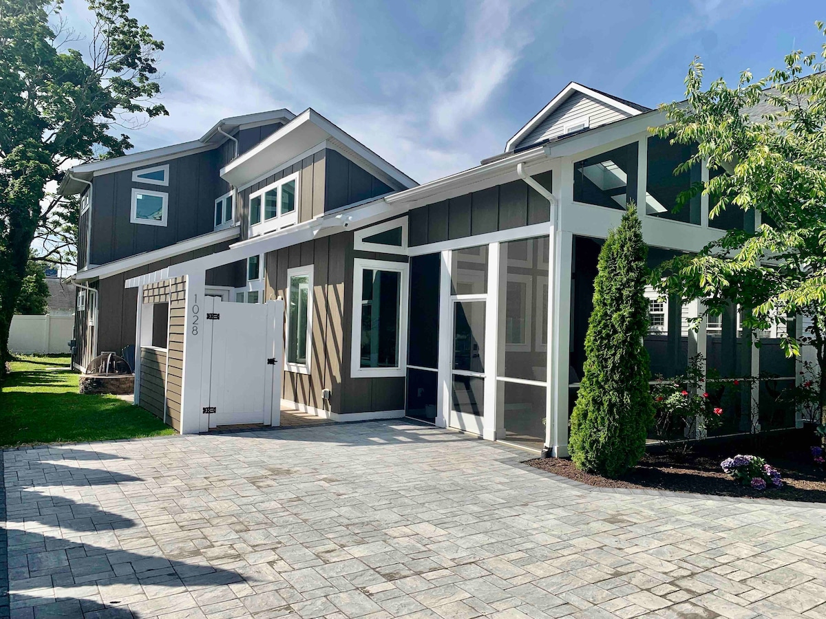 A modern beach home is showcased with a spacious paver driveway leading to both the main entrance and a gated area. The front features large windows and a screened porch, while greenery and landscaped beds frame the property, enhancing the inviting exterior.