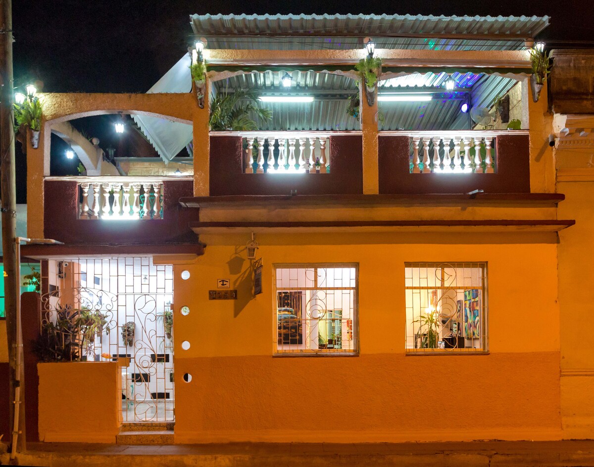 The exterior of a ground-floor apartment features a vibrant yellow façade with large windows, allowing views of the interior. The terrace above is adorned with plants and illuminated by soft lighting, enhancing its welcoming appearance at night.