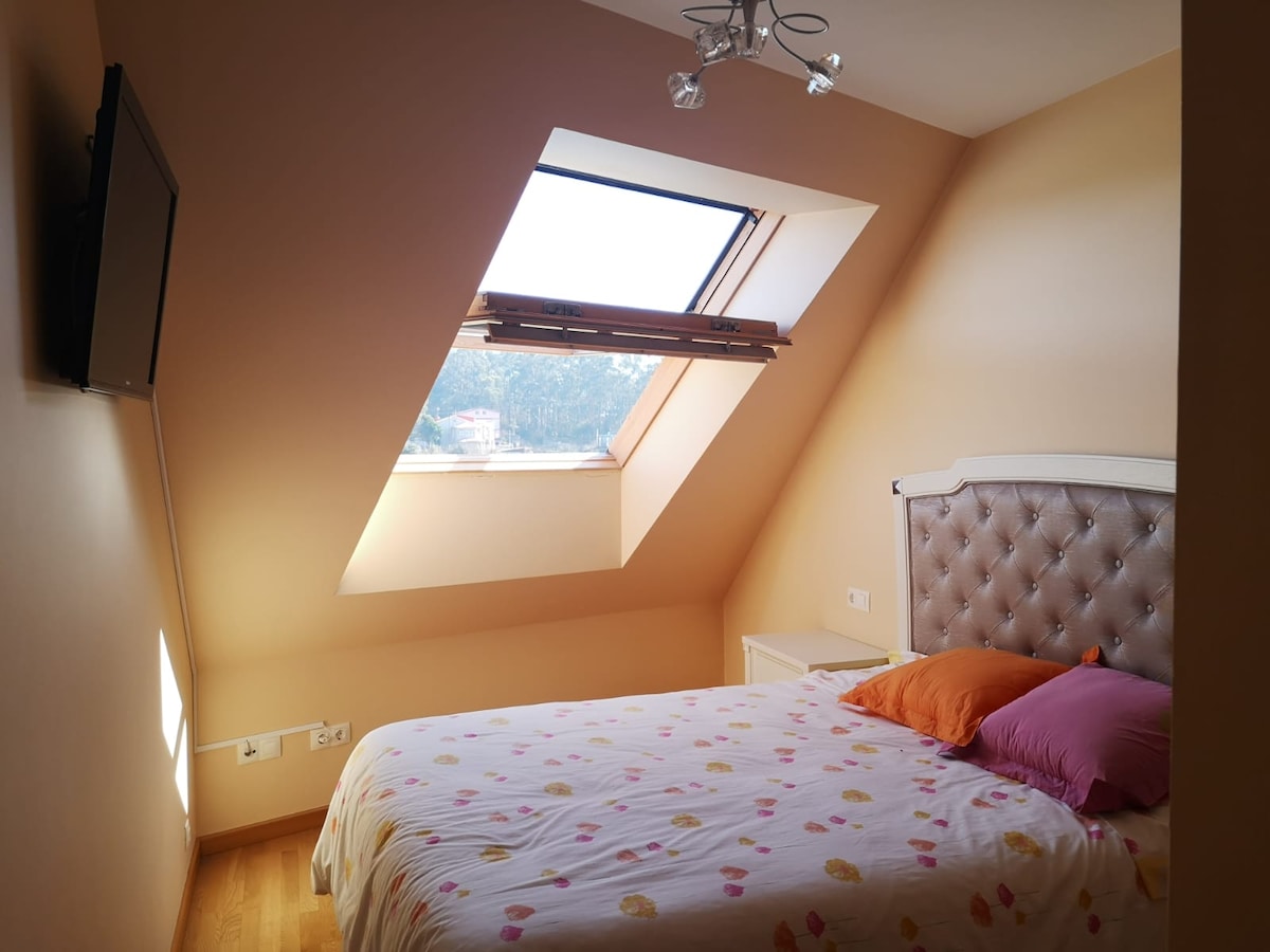 A cozy bedroom features a spacious bed with a floral-patterned duvet cover and pillows in shades of orange and purple. Natural light enters through a skylight, highlighting the beige walls and wooden floor. A mounted television is visible on one wall.
