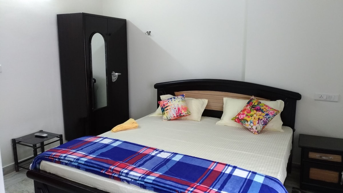 A spacious bed features a neutral-colored bedspread, complemented by decorative pillows with colorful patterns. A dark wooden wardrobe is positioned nearby, and a side table holds a small lamp. Natural light brightens the room, enhancing its comfortable appearance.
