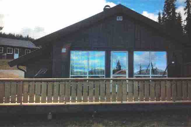 A wooden chalet features large windows reflecting the surrounding landscape. A spacious deck with a wooden railing offers outdoor seating possibilities. The structure’s sloped roof complements the natural setting, while trees and building elements are visible in the background.