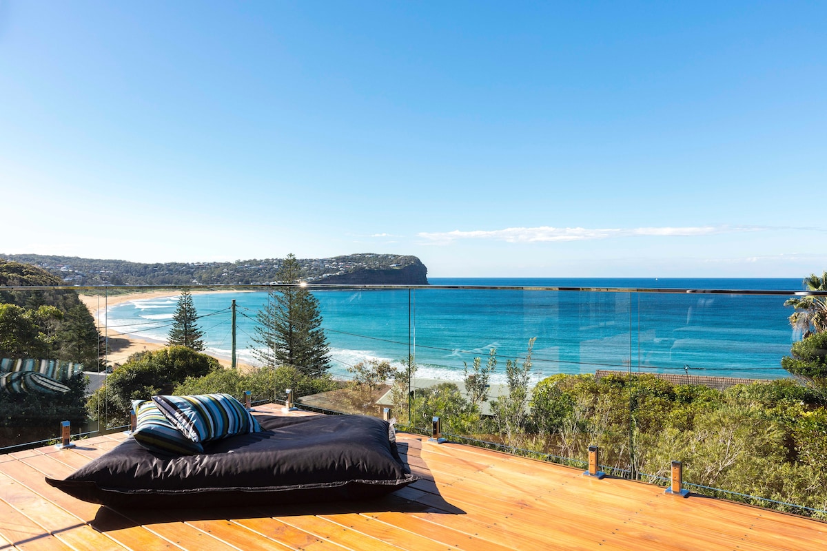 A spacious outdoor deck offers breathtaking ocean views stretching toward the horizon. A large black cushion sits on the wooden floor, inviting relaxation. Glass railings enhance the panoramic landscape while surrounding greenery adds natural beauty to the serene environment.