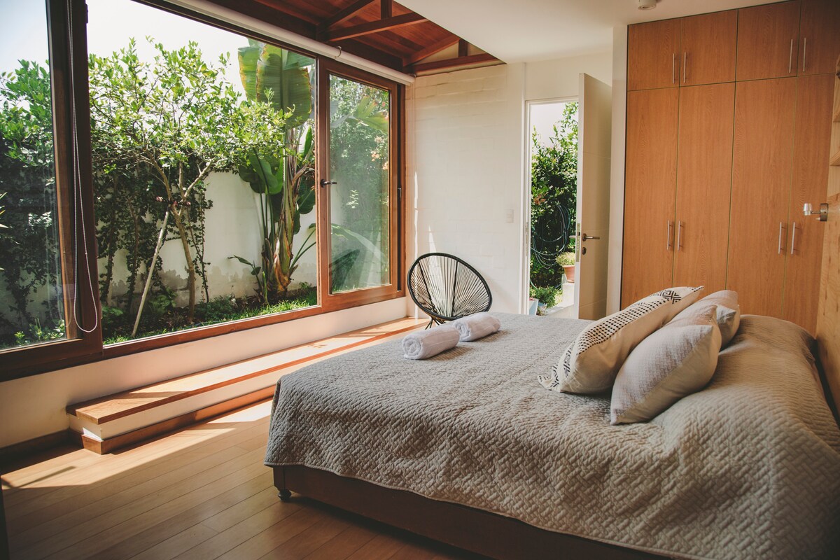 A bright bedroom features a large window with views of greenery. Light hardwood flooring complements the spacious bed, which is adorned with neutral-toned linens and pillows. A cozy chair is positioned nearby, and wooden cabinetry offers storage solutions.
