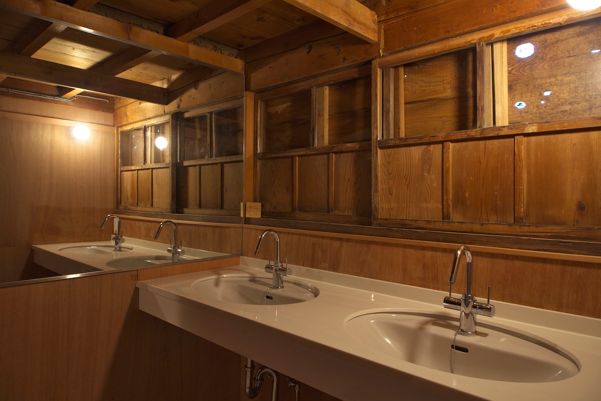 A shared bathroom area is illuminated by soft light, featuring two sinks mounted on a wooden counter. Natural materials are used throughout, with wooden walls and windows providing a rustic ambiance.