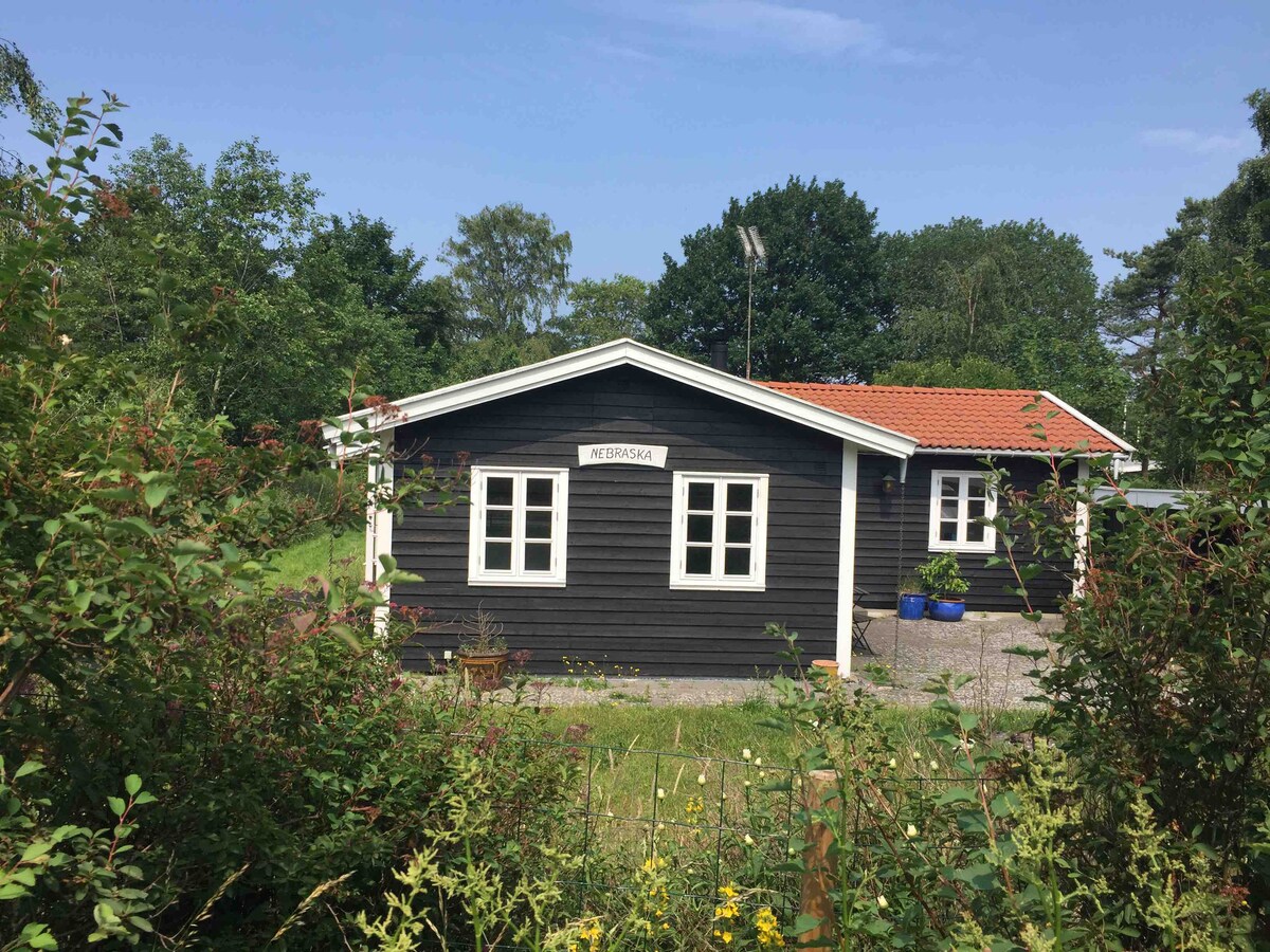 A single-story house is framed by lush greenery and flowering plants. The exterior is painted in dark tones with white window frames. A small sign indicates the house name. A gravel path leads to the entrance, surrounded by an inviting landscape.
