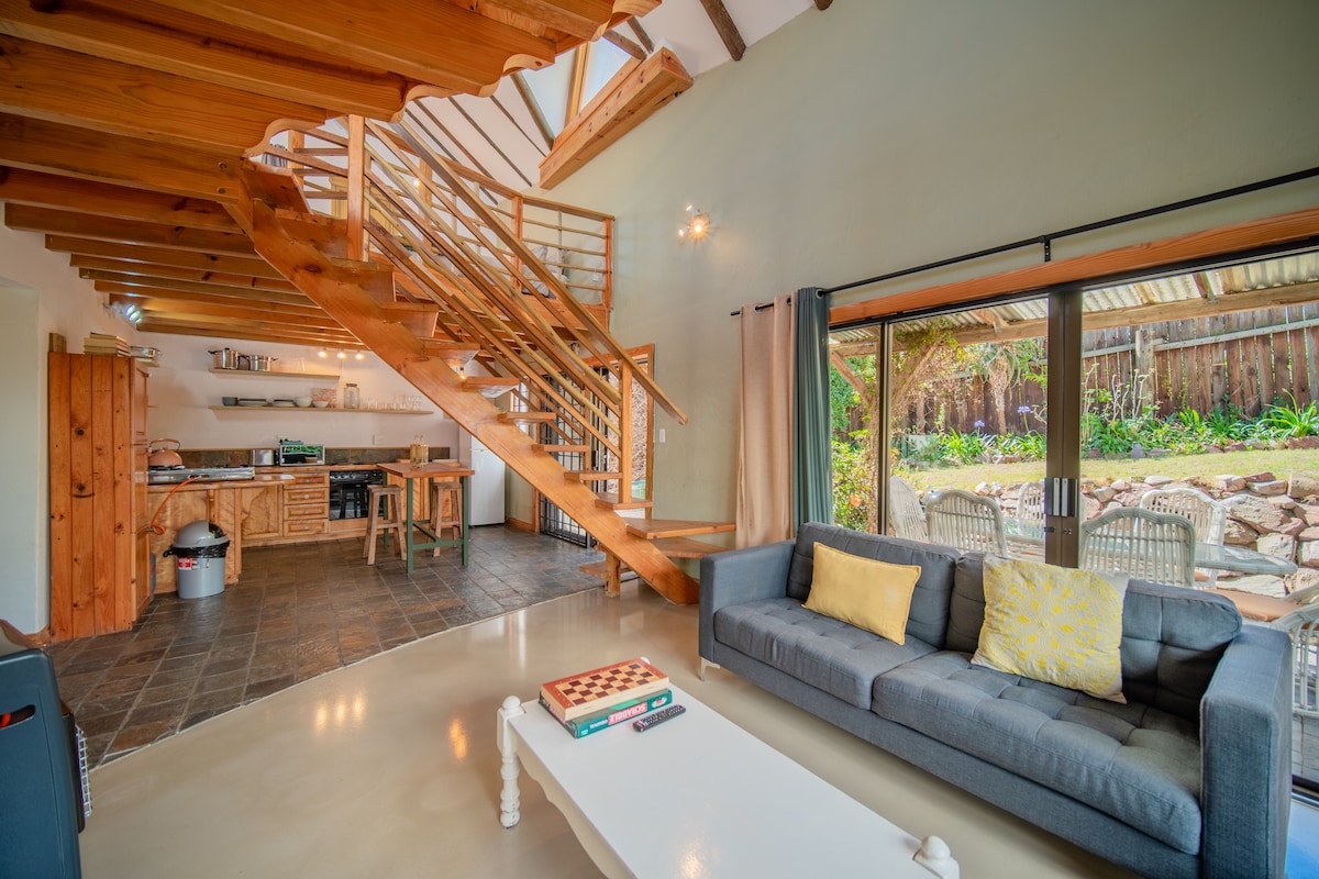 An open living area features a cozy lounge with a gray sofa, a white coffee table, and board games. A wooden staircase leads to the upper level. Large glass doors open to a private outdoor space, where seating is arranged amidst lush greenery.