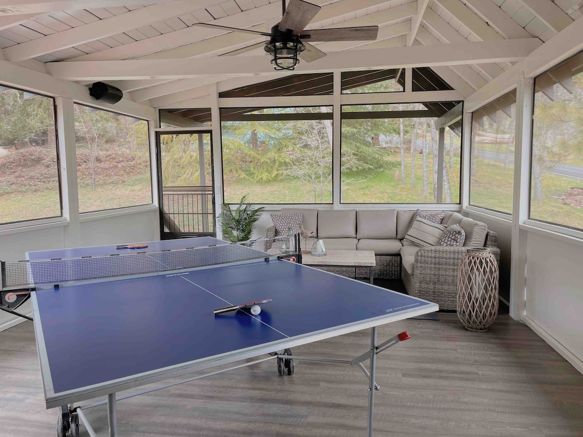 A screened-in room features a ping pong table in the center, surrounded by comfortable seating arrangements. Natural light enters through large windows, providing views of the outdoors. A ceiling fan hangs overhead, adding to the inviting space.