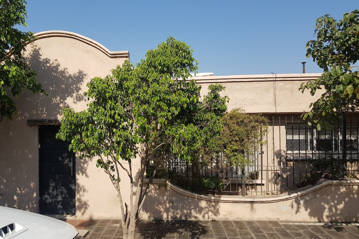 The exterior of a rustic home is framed by green foliage, featuring a small tree in front. The structure has a smooth beige facade with an arched top and a dark door, complemented by barred windows that allow natural light to enter.