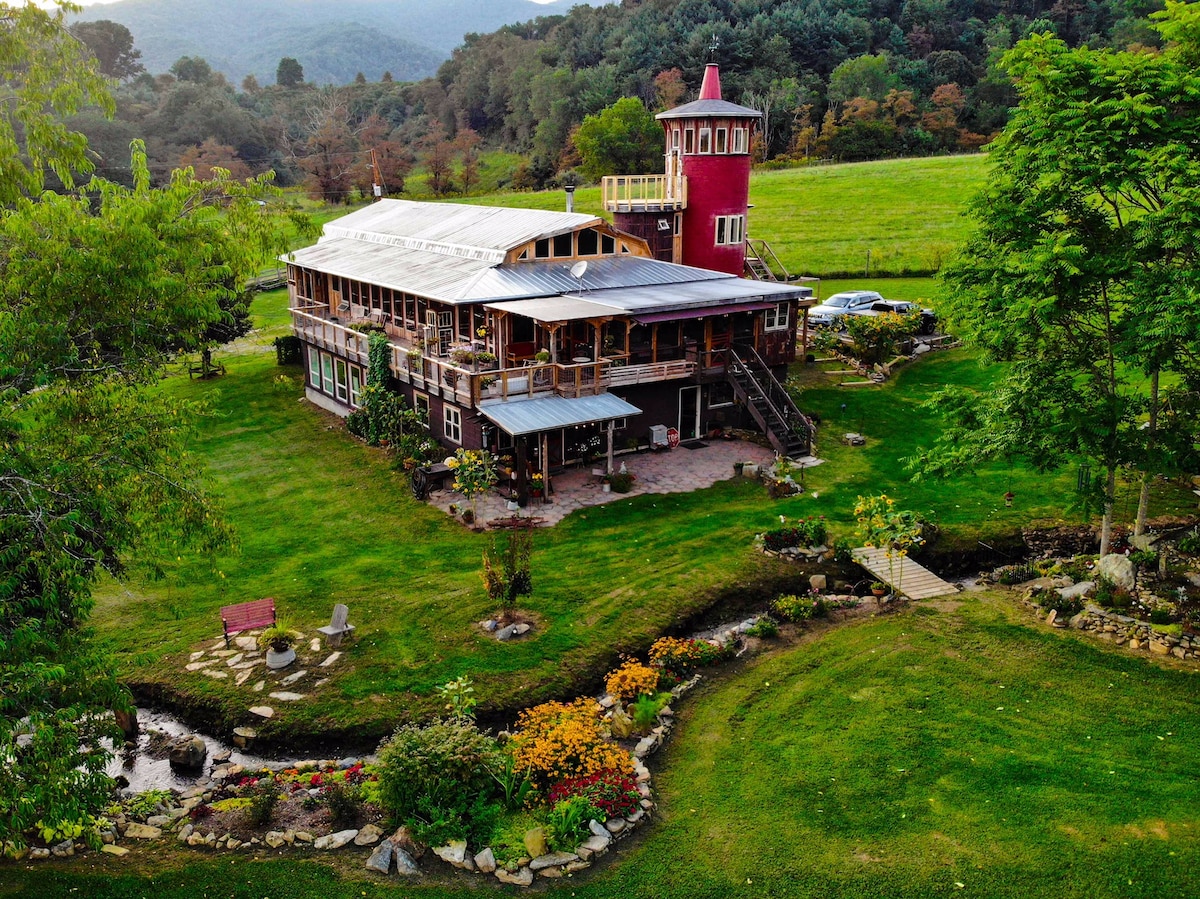 The rustic barn, set in a picturesque valley, features a multi-level structure with a distinctive red tower. Lush greenery surrounds the property, with a landscaped garden and stone pathways winding through vibrant flowers and a bubbling creek.