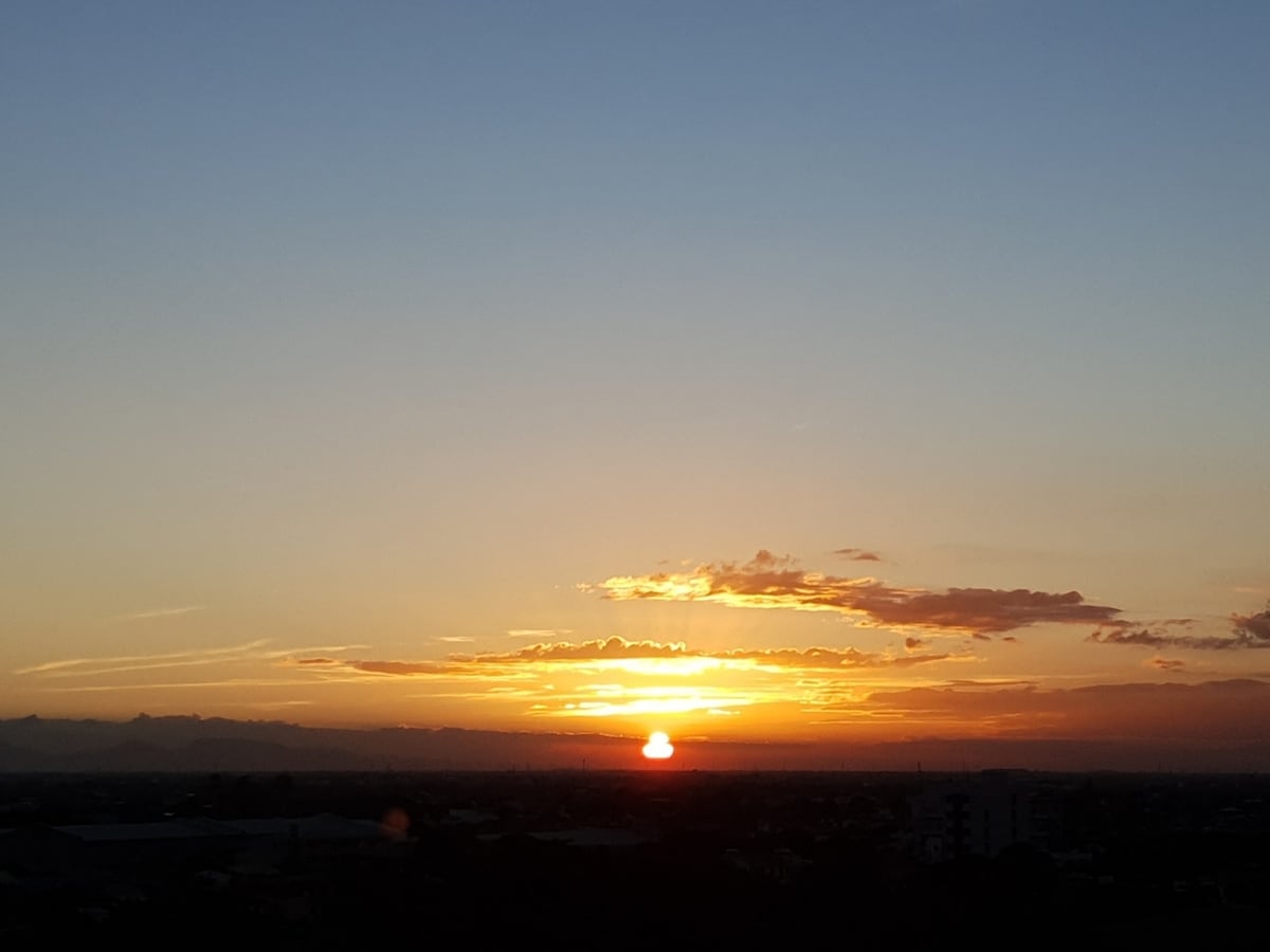 A sunset view is captured from the balcony, displaying vibrant orange and yellow hues against a clear sky. The horizon is marked by distant mountains, creating a peaceful contrast to the urban landscape below.