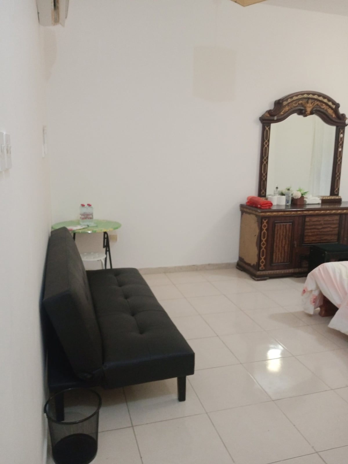 A comfortable seating area features a black sofa positioned against a white wall. A small round table with water bottles is located next to the sofa. A vintage-style dresser with a large mirror stands opposite, enhanced by neatly arranged toiletries and decorative items.