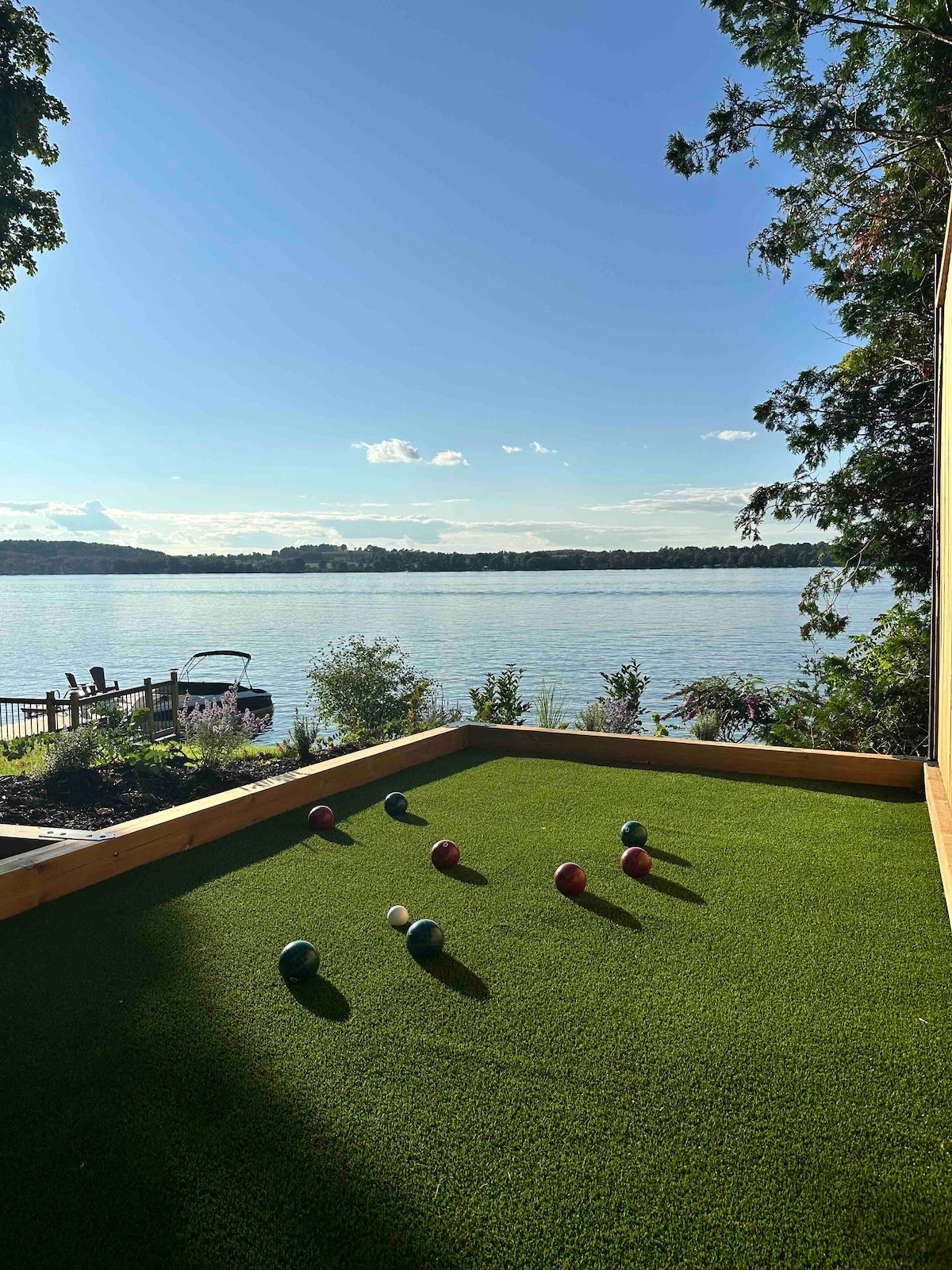 A bocce ball court is positioned against a serene lakeside backdrop, featuring bright green turf and colorful balls scattered across the playing area. The smooth water reflects the clear sky, with trees and a dock visible in the background.