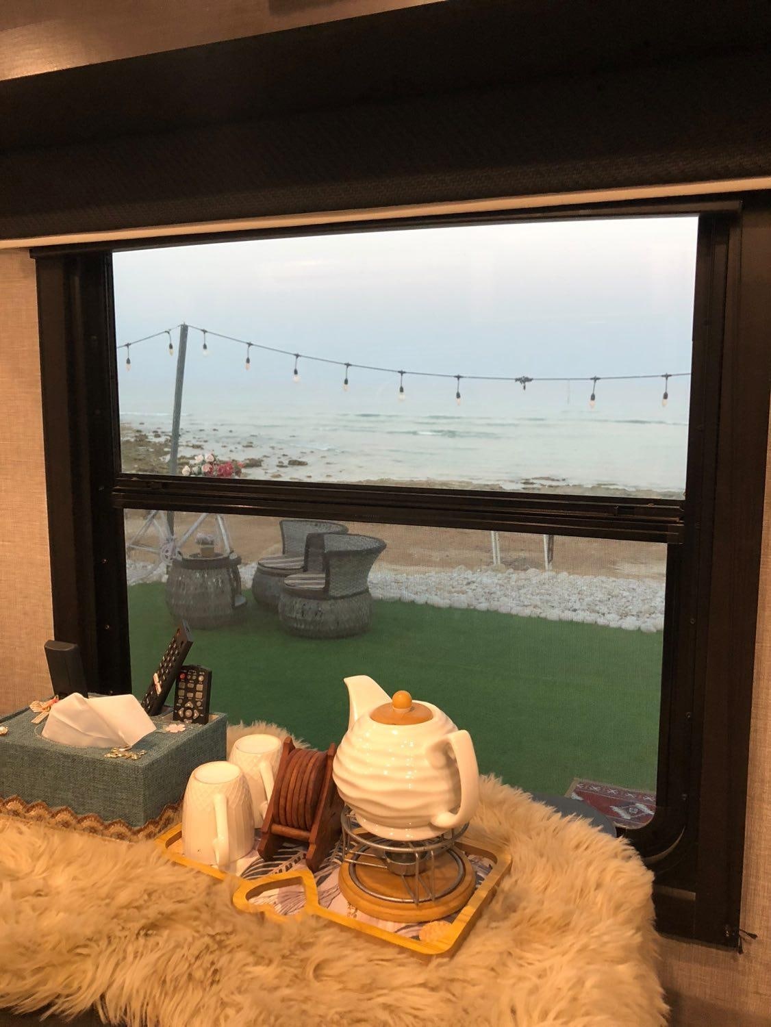 A window view frames the serene coastline, where gentle waves meet a sandy shore. Outdoor seating is visible, accompanied by decorative string lights overhead. A cozy interior space is suggested by the fur throw and various items arranged on a surface below the window.