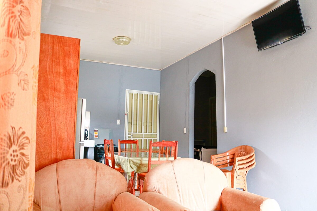 A living area features two comfortable, upholstered chairs facing each other. A television is mounted on the wall, while an archway leads to a dining space equipped with wooden chairs and a table. The room is illuminated by natural light from the adjacent area.