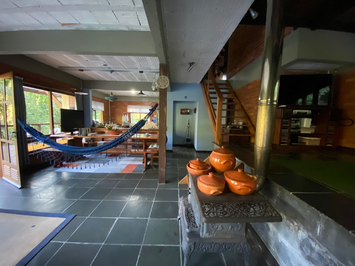 An open living space is shown, featuring large windows that allow natural light to fill the room. A hammock is hung for relaxation, and a dining area with wooden tables can be seen. Decorative clay pots are arranged on a side table, and stairs lead to another level.