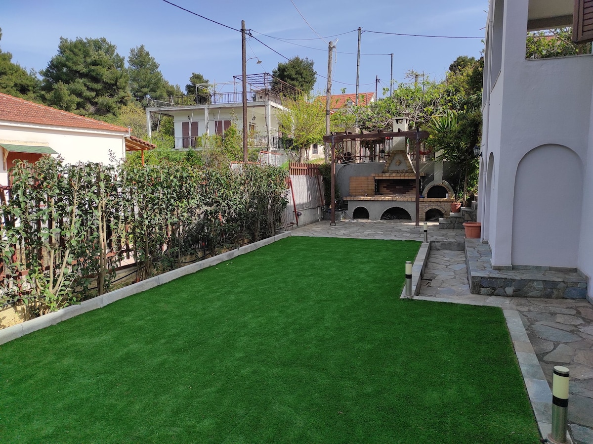 An outdoor area features a spacious garden with artificial grass, providing a vibrant green space for relaxation. A traditional stone oven and BBQ are present, surrounded by leafy plants and trees. The layout offers a clear path, enhancing the inviting atmosphere for gatherings.