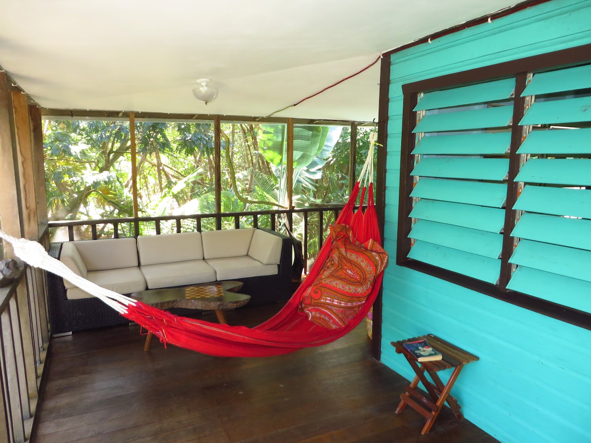 A comfortable seating area is featured on a screened-in porch, with a light-colored sofa and a vibrant red hammock. Natural light filters through the open windows, highlighting the tropical greenery outside. A small table and chair are also present, adding to the inviting space.