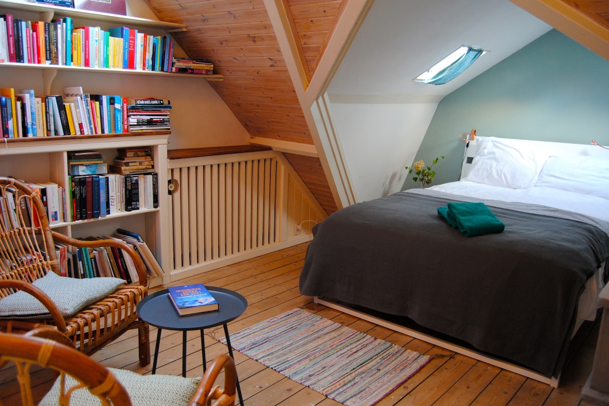 A cozy attic room features a spacious bed with a dark throw neatly arranged on top. A small round table and two woven chairs provide a reading nook. A bookshelf filled with various books lines the wall, and a skylight offers natural light.