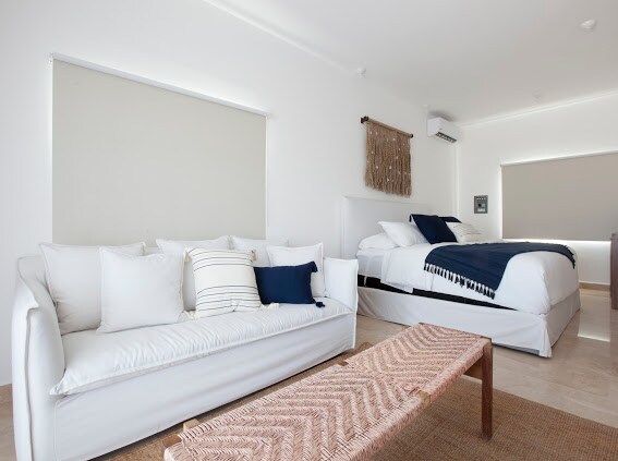 A light and airy bedroom space features a spacious bed covered with dark bedding and a decorative wall hanging. A white sofa adorned with various cushions provides additional seating, while a woven rug adds texture to the floor.