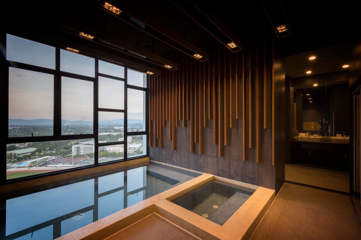 An indoor relaxation area features a sleek, modern design with a large window offering expansive views of the city and mountains. A luxurious soaking tub is positioned beside the glass, transforming the space into a tranquil retreat illuminated by warm lighting.
