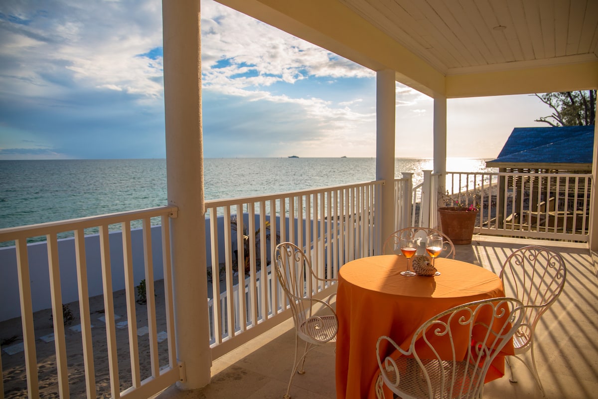 A covered outdoor balcony features a round table dressed in orange cloth, surrounded by white wrought iron chairs. The ocean view is visible, with gentle waves and a colorful sky at sunset providing a serene backdrop.