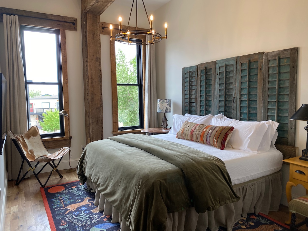 The room features a spacious bed with a muted green throw blanket, complemented by decorative pillows. Natural light filters in through two large windows, enhancing the rustic charm of the wooden shutter headboard and exposed concrete accents. A small lamp and a chair add functional decor.