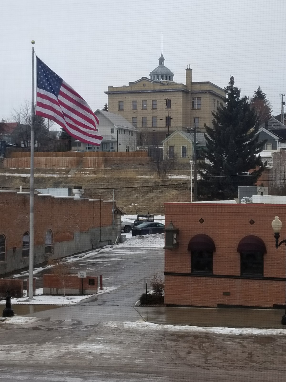 An American flag flutters gently in the foreground, with a view of a historic building in the background. The scene features a mix of snow and bare ground, with a few parked vehicles visible along the street.