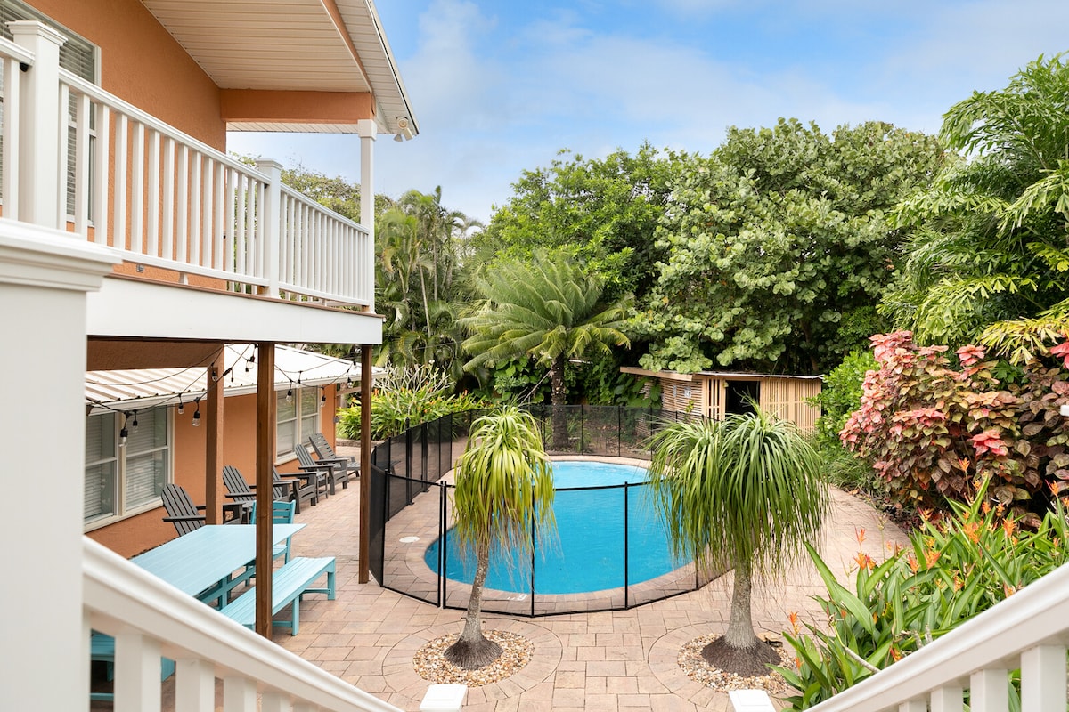 A tranquil backyard oasis features a sparkling pool surrounded by lush greenery. Lounge chairs are positioned around the pool area, with a safety fence enclosing it. Palm trees and vibrant plants add to the serene atmosphere, framed by the home's balcony above.