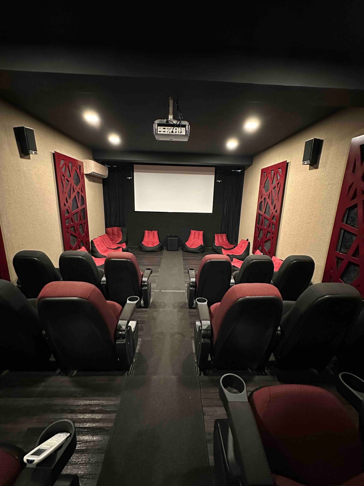A private screening room is equipped with comfortable seating arranged in tiered rows. The space features a large projector screen at the front, flanked by sound speakers and illuminated by soft overhead lights, creating an ideal environment for movie viewing.