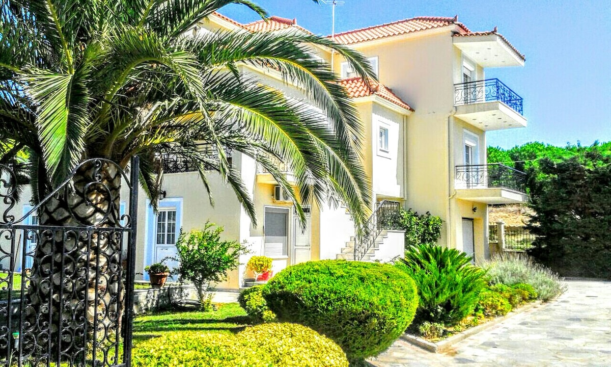 The exterior of a three-story building is shown, featuring a blend of light-colored walls and tiled roofs. Lush greenery surrounds the entrance, including well-maintained shrubs and palm trees. A gated pathway leads to the front door, enhancing the inviting appeal of the property.