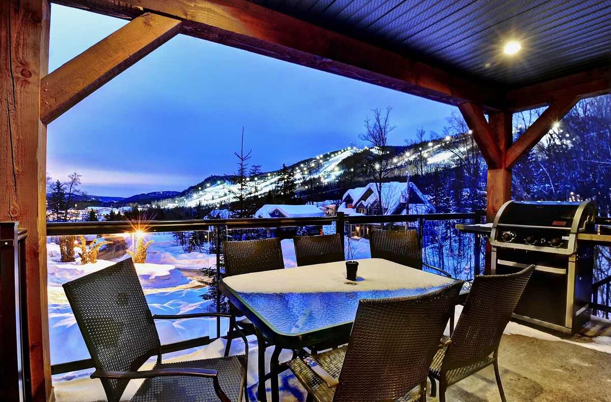 An outdoor deck is featured with a spacious table surrounded by several chairs. A barbecue grill is positioned nearby. In the background, snow-covered slopes are visible, illuminated by lights from the nearby ski area, enhancing the mountain view.