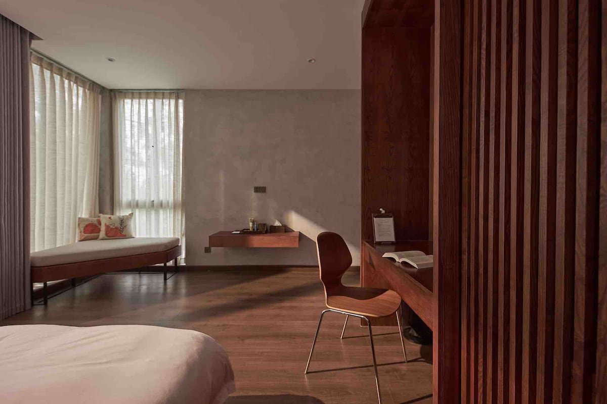 A spacious room features large windows that allow natural light to fill the space. A comfortable bed is positioned next to a modern wooden desk with a chair. A cozy seating area is present by the window, with a simple bench adorned with decorative cushions.