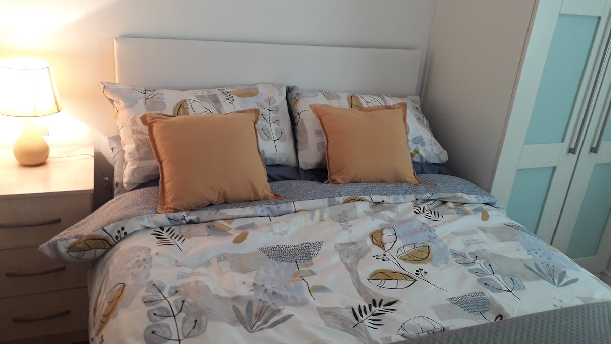 A neatly arranged sleeping area features a spacious bed dressed in a patterned duvet cover with soft, neutral tones. Two decorative pillows add visual interest. A bedside lamp provides gentle illumination, and a storage unit is visible to the left, offering additional functionality.