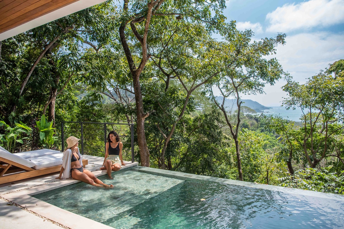 A tranquil infinity pool sits on a sunlit terrace, overlooking the lush greenery and ocean in the distance. Two individuals relax by the water's edge, enjoying the serene environment. Sun loungers are positioned nearby, surrounded by tropical foliage and expansive views.