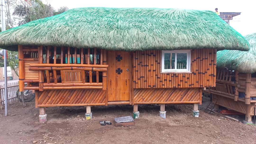 A charming bamboo cottage features a thatched roof and wooden accents. The front porch is equipped with seating, while a small white window allows natural light into the interior. The entrance door is adorned with decorative elements, enhancing the rustic appeal of the structure.