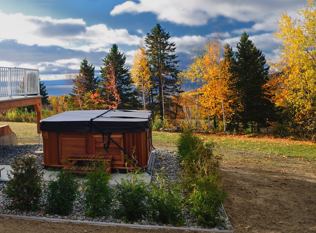 An outdoor hot tub is surrounded by landscaped greenery and vibrant autumn foliage. The view extends to the distant mountains and body of water under a partly cloudy sky, creating a serene and inviting environment.